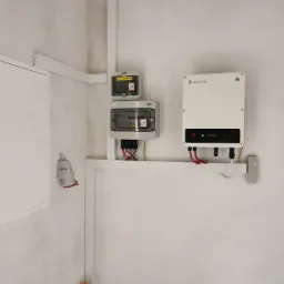 Biały inwerter GoodWe zamontowany na ścianie obok dwóch skrzynek elektrycznych KENO, z widocznymi przewodami i elementami instalacji, na tle surowej, jasnej ściany.