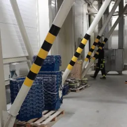 Wnętrze hali przemysłowej z białymi, ukośnymi słupami pomalowanymi w czarno-żółte pasy ostrzegawcze, ułożone niebieskie pojemniki na paletach, pracownik w uniformie w tle.