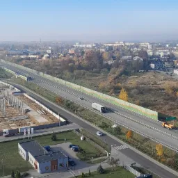 Widok z góry na budynek biurowy z parkingiem, fragment autostrady z ekranami akustycznymi i billboard reklamowy z logo Sievert na trawniku, w tle zabudowa miejska pod błękitnym niebem.