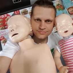 ZIELMED PAWEŁ ZIELONKA - Instruktor z dwoma manekinami do nauki RKO niemowląt na tle menu fast food. Uśmiechnięty mężczyzna prezentuje sprzęt szkoleniowy.