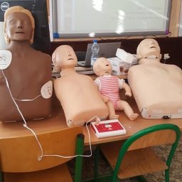 ZIELMED PAWEŁ ZIELONKA - Manekiny do ćwiczeń RKO na biurku w sali lekcyjnej, w tle tablica interaktywna z prezentacją o pierwszej pomocy. Jeden z manekinów ma elektrody EKG.