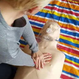 Instruktorka demonstruje masaż serca na manekinie z osłoną twarzy, leżącym na kolorowym kocu w paski, podczas praktycznego szkolenia z pierwszej pomocy.