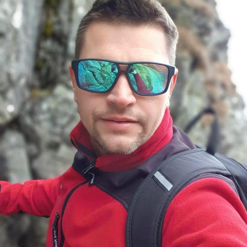 Mężczyzna w czerwonej polarowej kurtce i okularach przeciwsłonecznych z odbiciem górskiego krajobrazu, sugerujący aktywność na świeżym powietrzu i potencjalne szkolenie outdoor.