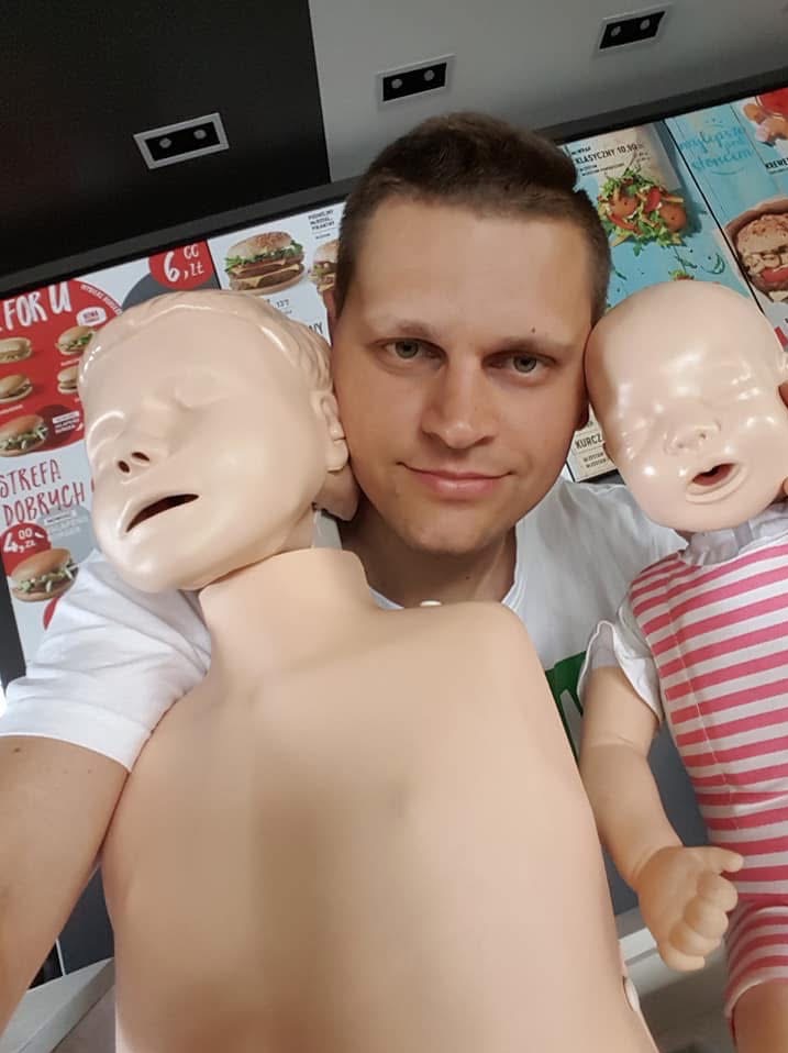 Instruktor z dwoma manekinami do nauki RKO niemowląt na tle menu fast food. Uśmiechnięty mężczyzna prezentuje sprzęt szkoleniowy.