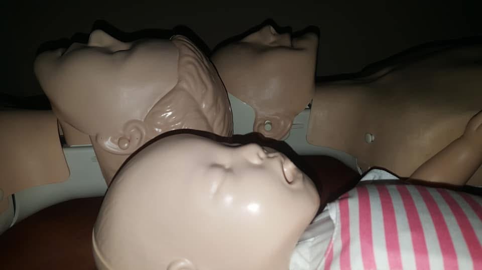 Manekiny do nauki pierwszej pomocy, w tym jeden manekin niemowlęcia w bluzie w biało-różowe paski, w słabym oświetleniu.