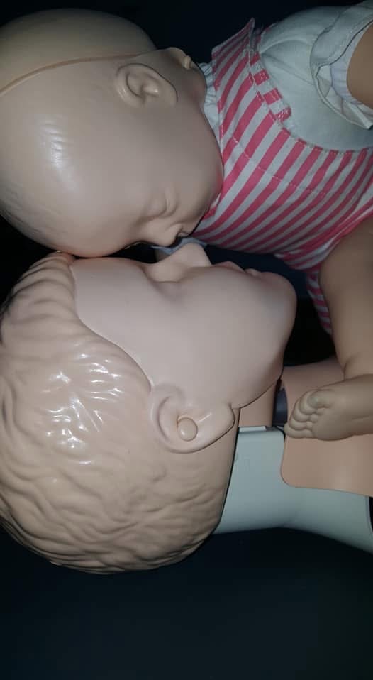 Manekiny do nauki RKO: dorosły i niemowlę, widoczne z bliska. Realistyczne włosy u dorosłego, różowa bluzka w paski u niemowlaka, ciemne tło.