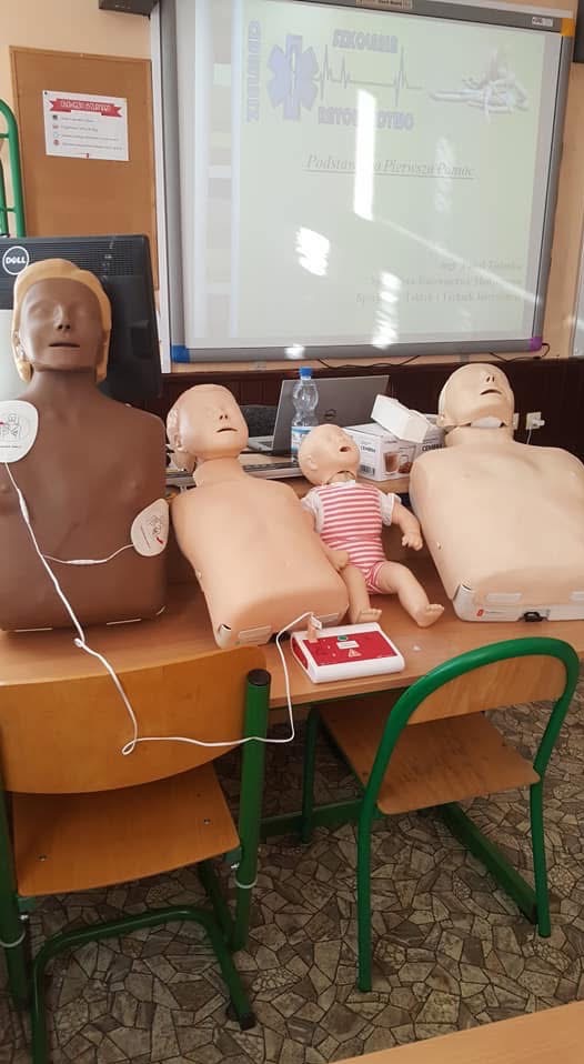 Manekiny do ćwiczeń RKO na biurku w sali lekcyjnej, w tle tablica interaktywna z prezentacją o pierwszej pomocy. Jeden z manekinów ma elektrody EKG.