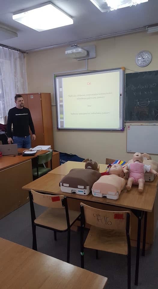 Sala szkoleniowa z manekinami do ćwiczeń resuscytacji, projektor wyświetla slajd z celem kursu, prowadzący stoi obok biurka z laptopem.