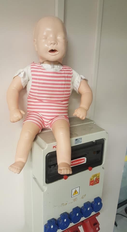 Manekin niemowlęcia w różowo-białym ubranku siedzi na białej skrzynce z gniazdami. Scena z kursu pierwszej pomocy dla niemowląt.