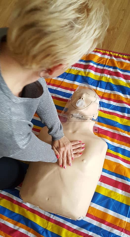 Instruktorka demonstruje masaż serca na manekinie z osłoną twarzy, leżącym na kolorowym kocu w paski, podczas praktycznego szkolenia z pierwszej pomocy.