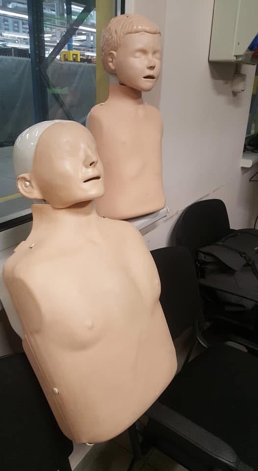 Dwa fantomy do nauki RKO: manekin z białą głową na krześle i drugi z włosami w tle, w pomieszczeniu z elementami instalacji przemysłowej.