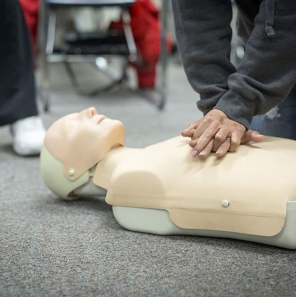 Instruktor demonstruje resuscytację krążeniowo-oddechową na manekinie. Dłonie ułożone na klatce piersiowej, widoczne detale stroju, sala szkoleniowa w tle.