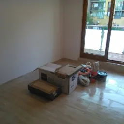 Przygotowania do malowania: puszki z farbami Dekoral i Dulux, kartony z ceramiką sanitarną, akcesoria malarskie w rogu jasnego pokoju z dużym oknem balkonowym i beżową podłogą z płytek.