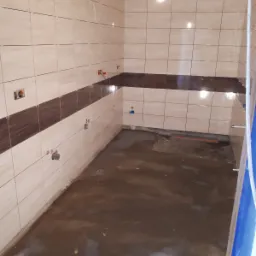 Wnętrze w trakcie prac wykończeniowych: ściany wyłożone beżowymi płytkami z brązowym pasem, widoczne wystające rurki instalacji, podłoga z wylewką w trakcie schnięcia.