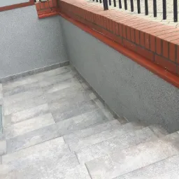 Zewnętrzne schody wyłożone szarymi płytkami prowadzące w dół, ściany wykończone szarym tynkiem strukturalnym, ceglany murek z czarną balustradą w tle.