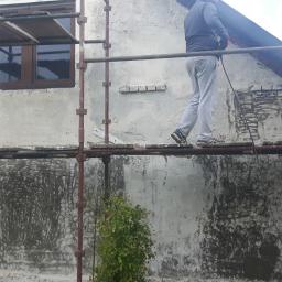 Adipat - Pracownik na rusztowaniu nanosi tynk na elewację domu jednorodzinnego, widoczne ślady po wcześniejszej warstwie, w tle fragment ogrodu.