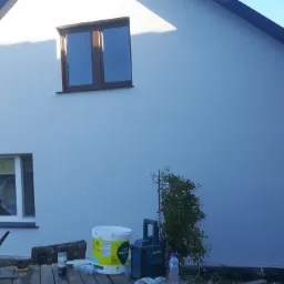 Świeżo pomalowana, jasnoszara elewacja domu jednorodzinnego z brązowymi ramami okiennymi, widoczne wiadro z farbą silikonową, narzędzia malarskie i pracownik układający kostkę brukową.