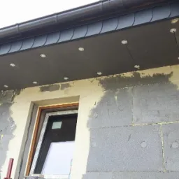 Fragment elewacji domu w trakcie ocieplania, widoczne płyty styropianowe z kołkami mocującymi oraz obróbka okienna przed nałożeniem siatki.