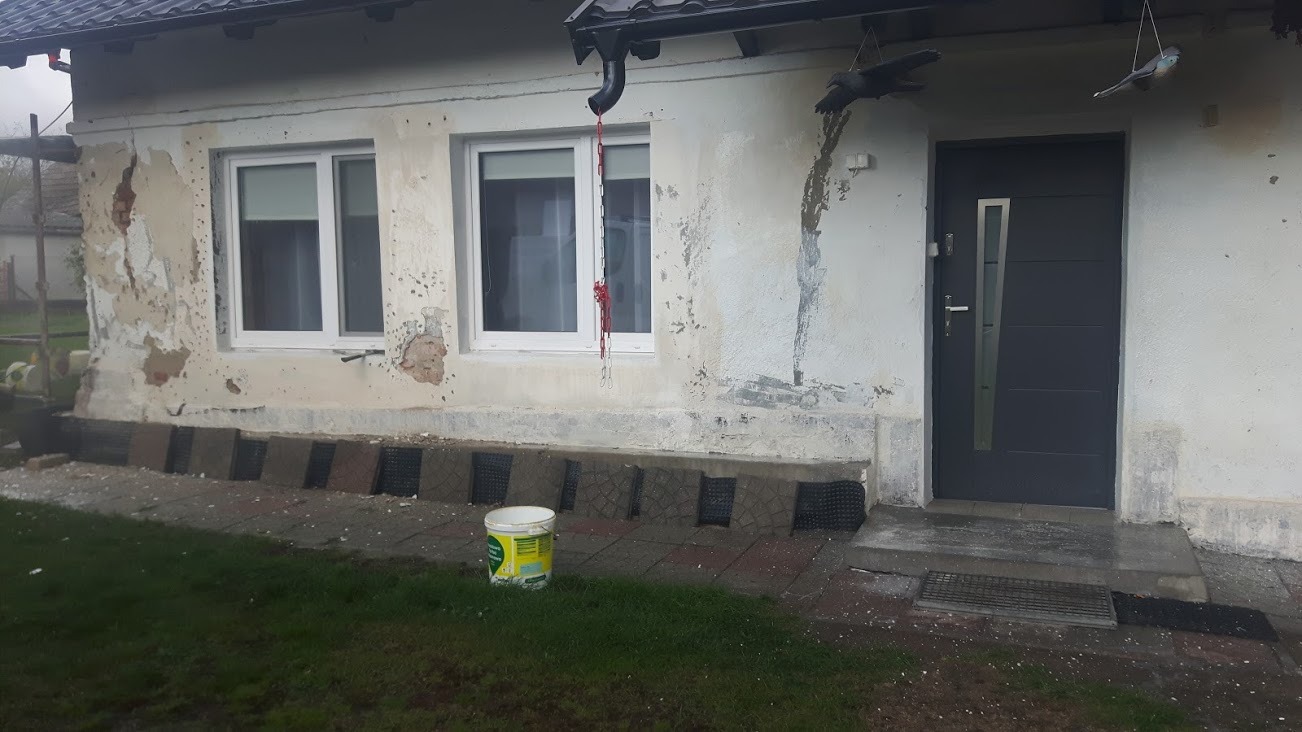 Niedokończona elewacja domu z widocznymi uszkodzeniami tynku, fragmentami odsłoniętej cegły i nowymi oknami; przed budynkiem ustawione płyty chodnikowe i wiadro z farbą.