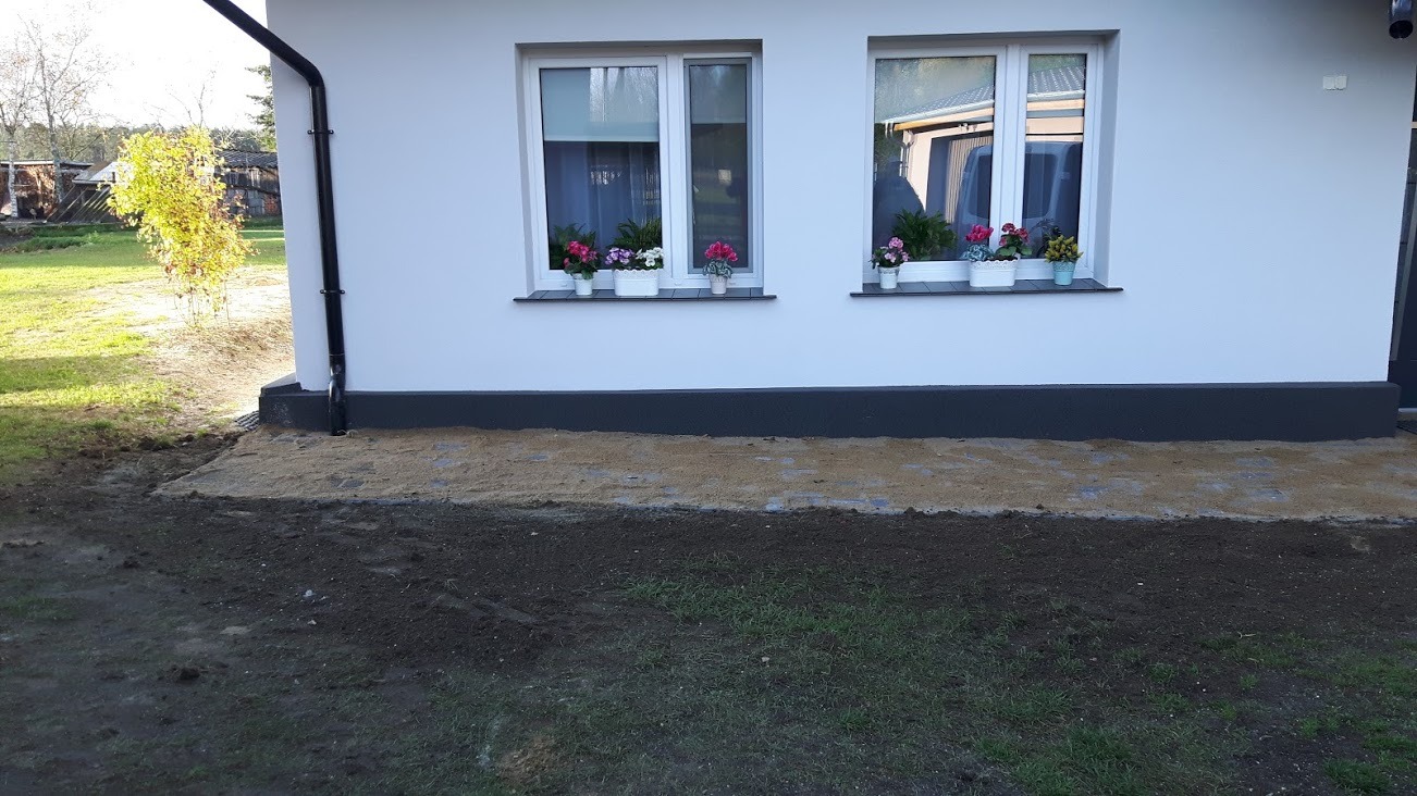 Biała elewacja domu z dwoma oknami ozdobionymi kwiatami w doniczkach, widoczny fragment obłożony piaskiem i częściowo ułożona kostka brukowa przy fundamencie budynku, trawnik i drzewa w tle.