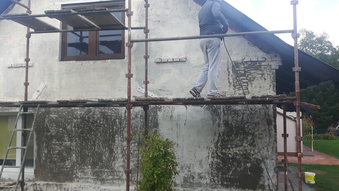 Pracownik na rusztowaniu nanosi tynk na elewację domu jednorodzinnego, widoczne ślady po wcześniejszej warstwie, w tle fragment ogrodu.