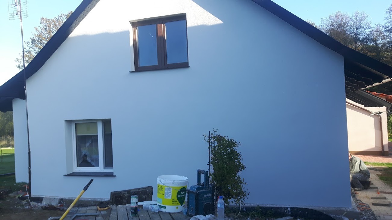 Świeżo pomalowana, jasnoszara elewacja domu jednorodzinnego z brązowymi ramami okiennymi, widoczne wiadro z farbą silikonową, narzędzia malarskie i pracownik układający kostkę brukową.