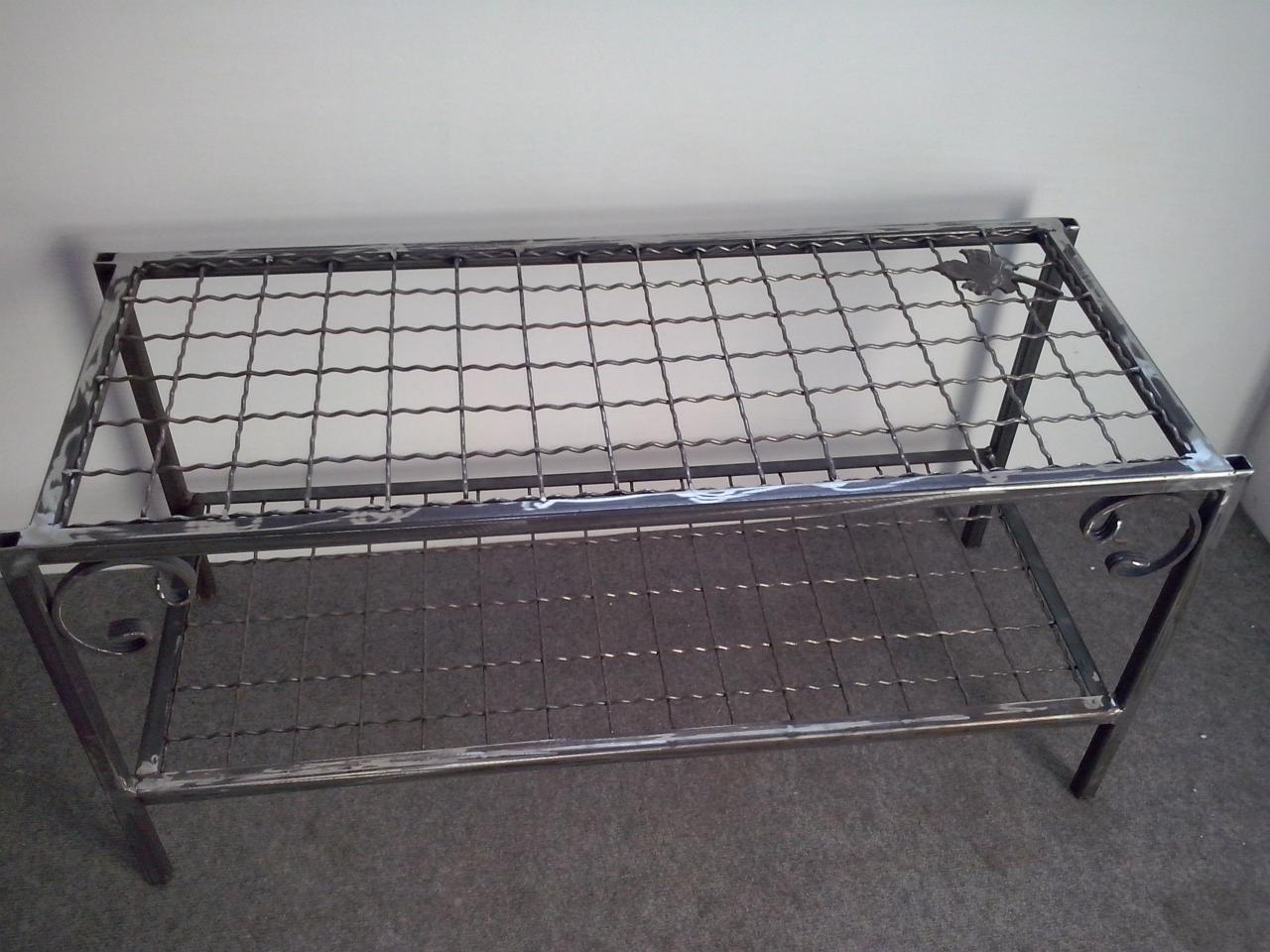 Metalowa konstrukcja regału z siatkowymi półkami, spawana z surowego metalu, dekoracyjne zawijasy na bokach, widoczne ślady spawania.