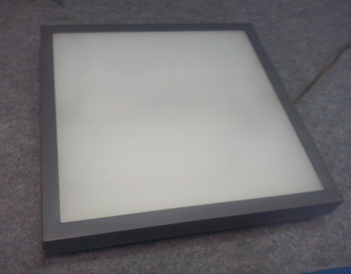 Kwadratowy panel LED z szarą ramą, leżący na szarej powierzchni, widoczny przewód zasilający w prawym górnym rogu.