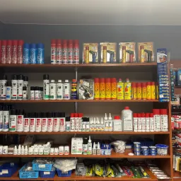 Półki w sklepie z elektroniką wypełnione różnorodnymi środkami czyszczącymi, smarami, klejami, lutami i bateriami, w tym spraye Kontakt, WD-40 i baterie Panasonic, uporządkowane tematycznie.