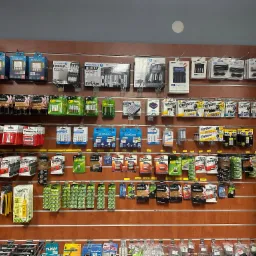 Ekspozycja w sklepie z szerokim wyborem baterii i ładowarek różnych typów i marek, wiszących na panelu ściennym z poziomymi szczelinami.