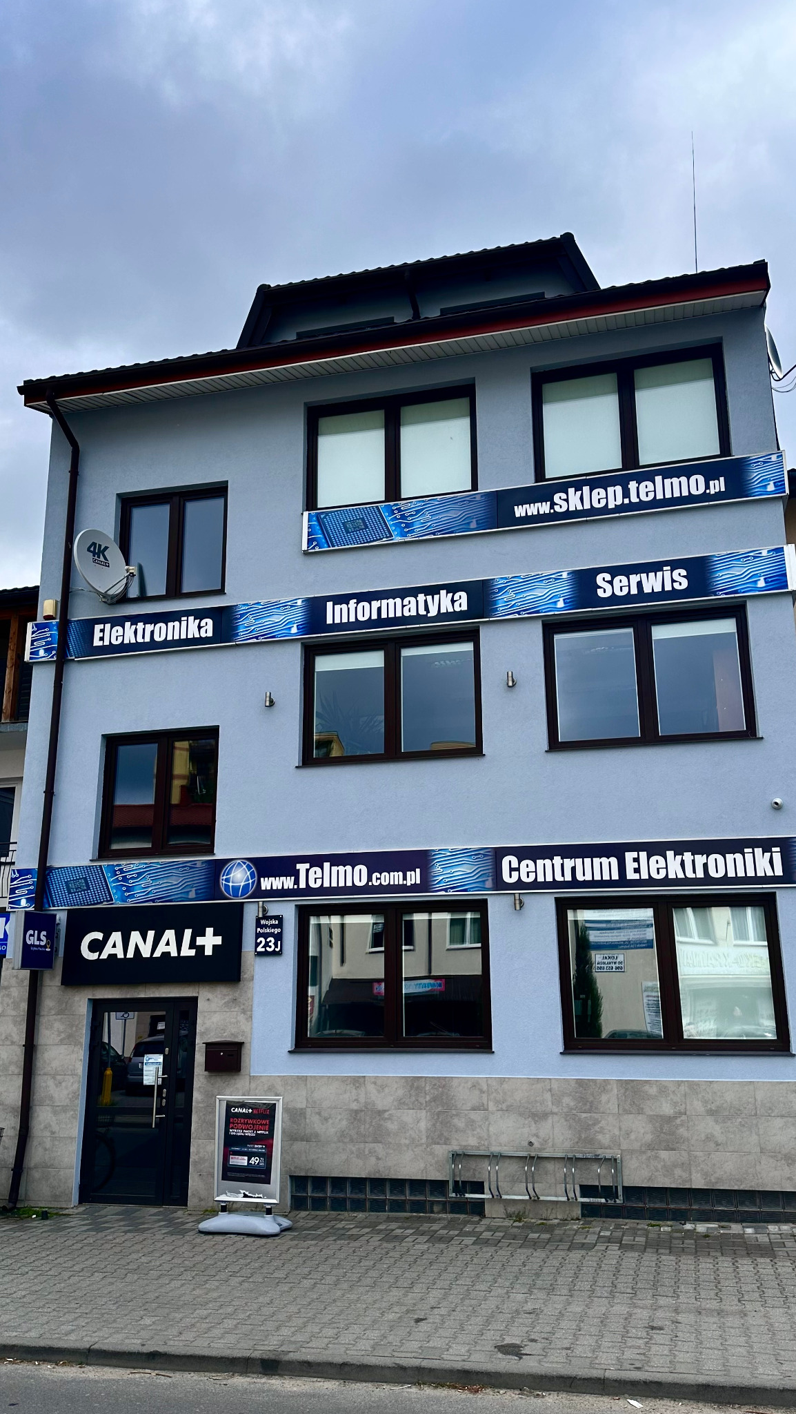 Elewacja budynku z szyldami informującymi o działalności: Elektronika, Informatyka, Serwis, Centrum Elektroniki. Dodatkowo widoczne logo Canal+ oraz GLS.