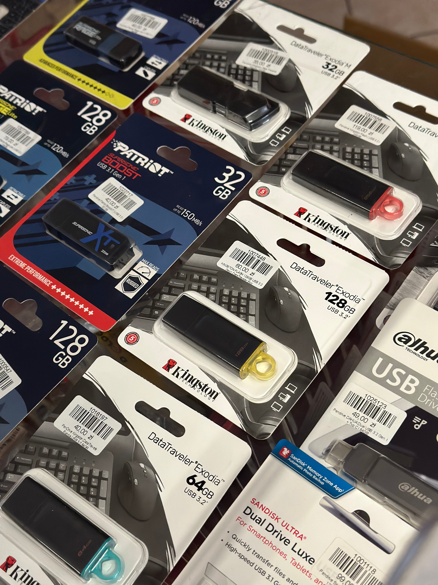 Różnorodność pendrive'ów USB różnych marek (Kingston, Patriot, SanDisk, Dahua) o pojemnościach od 32GB do 128GB, zapakowanych w plastikowe blistry z widocznymi cenami i specyfikacjami, ułożonych...