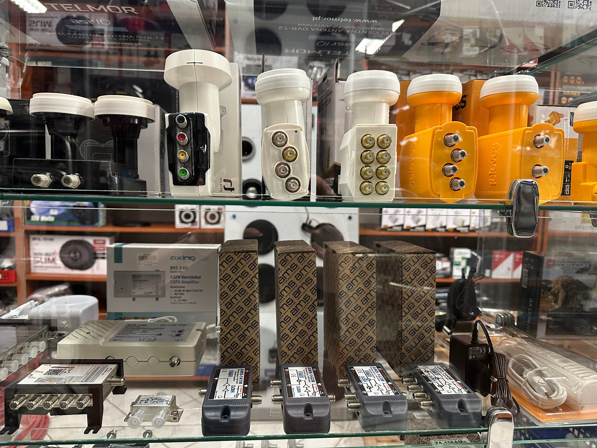 Różnokolorowe konwertery satelitarne i wzmacniacze CATV na szklanej półce w sklepie, widoczne pudełka z logo AMS i Televes.