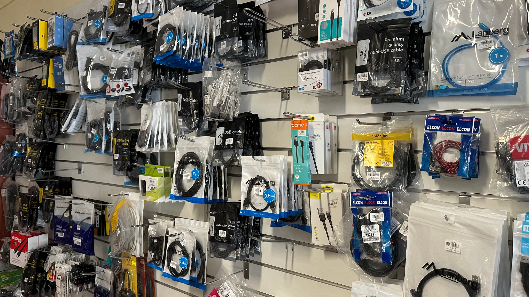 Ściana w sklepie z akcesoriami komputerowymi, pełna kabli USB, HDMI, przedłużaczy i adapterów, zapakowanych w foliowe opakowania i zawieszonych na metalowych listwach.