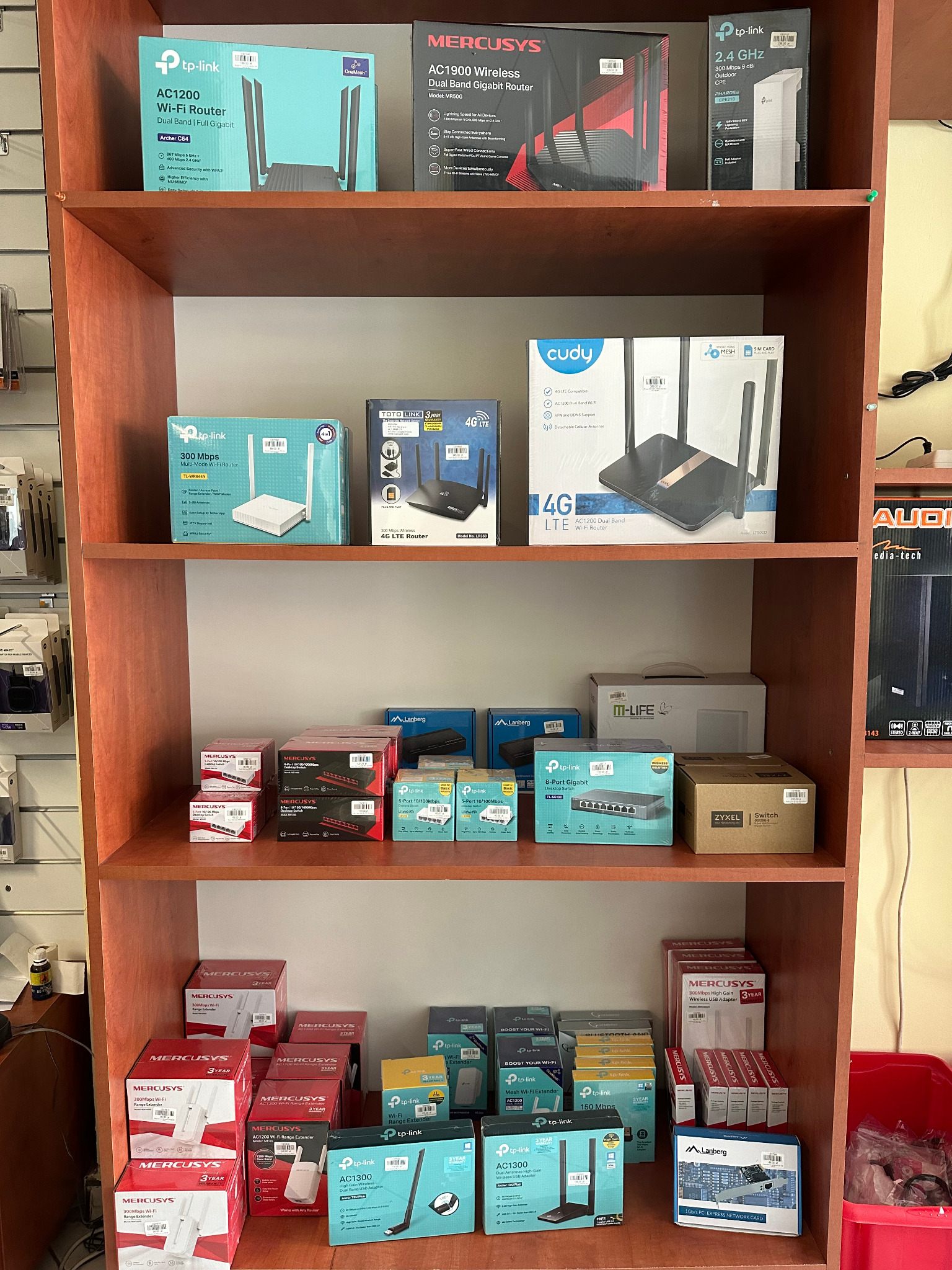 Półka sklepowa wypełniona pudełkami z routerami WiFi, switchami sieciowymi i kartami sieciowymi różnych producentów, takich jak TP-Link, Mercusys, Lanberg i Cudy.