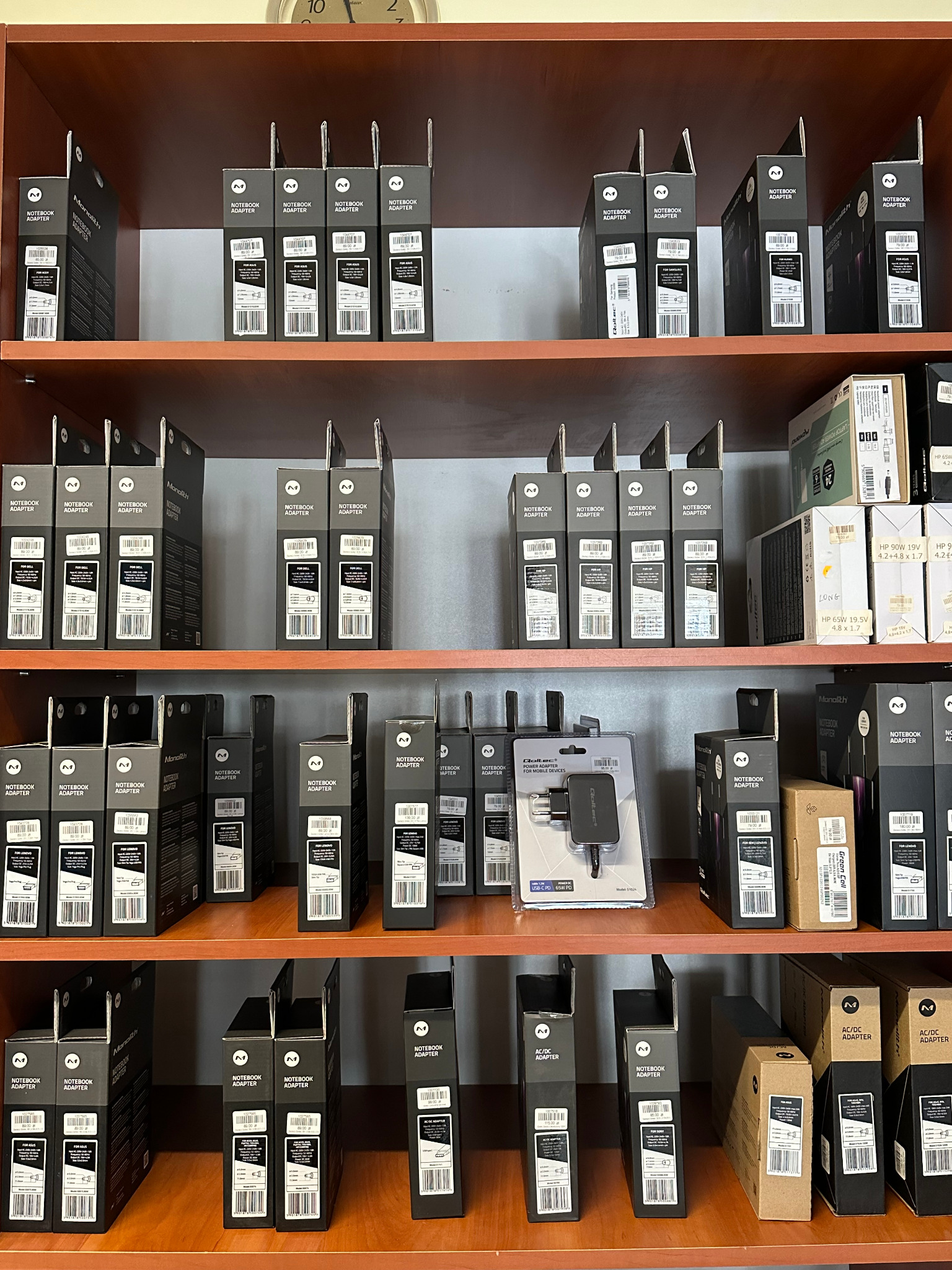 Półki w sklepie z akcesoriami komputerowymi, wypełnione pudełkami z zasilaczami do laptopów różnych marek, w tym Monolith i Qoltec, oraz adapterami USB-C.