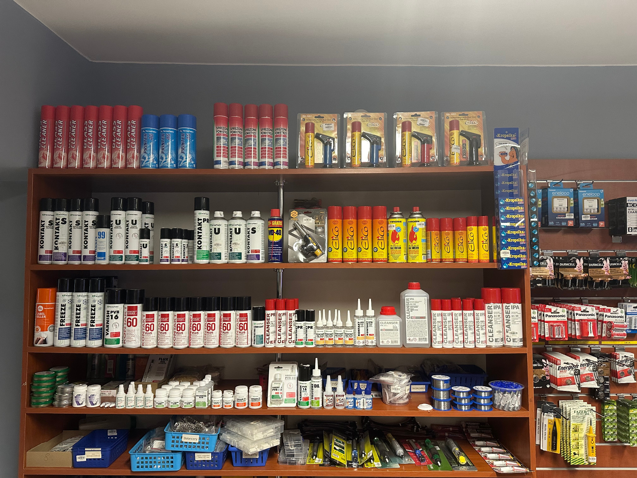 Półki w sklepie z elektroniką wypełnione różnorodnymi środkami czyszczącymi, smarami, klejami, lutami i bateriami, w tym spraye Kontakt, WD-40 i baterie Panasonic, uporządkowane tematycznie.