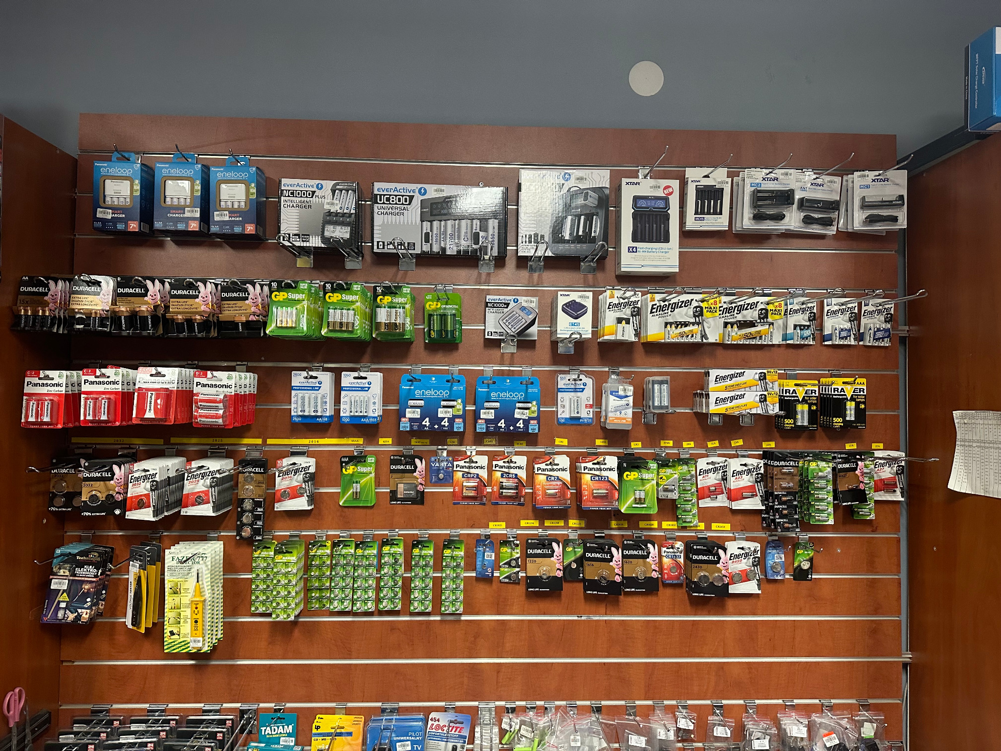Ekspozycja w sklepie z szerokim wyborem baterii i ładowarek różnych typów i marek, wiszących na panelu ściennym z poziomymi szczelinami.