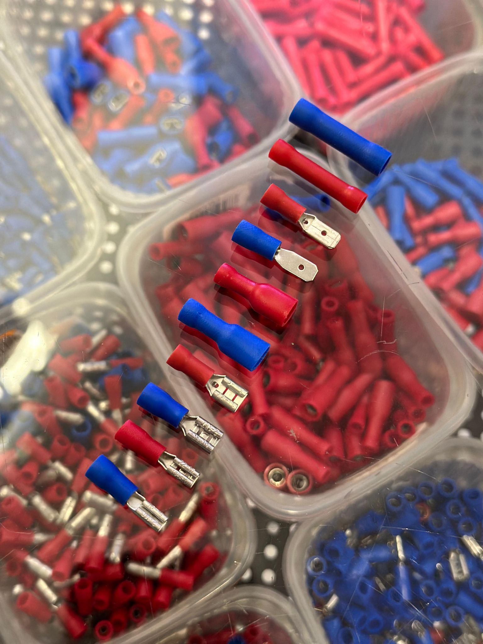 Zestaw przezroczystych pojemników z czerwonymi i niebieskimi tulejkami kablowymi, niektóre z metalowymi końcówkami, ułożone na blacie z wzorem w kropki.