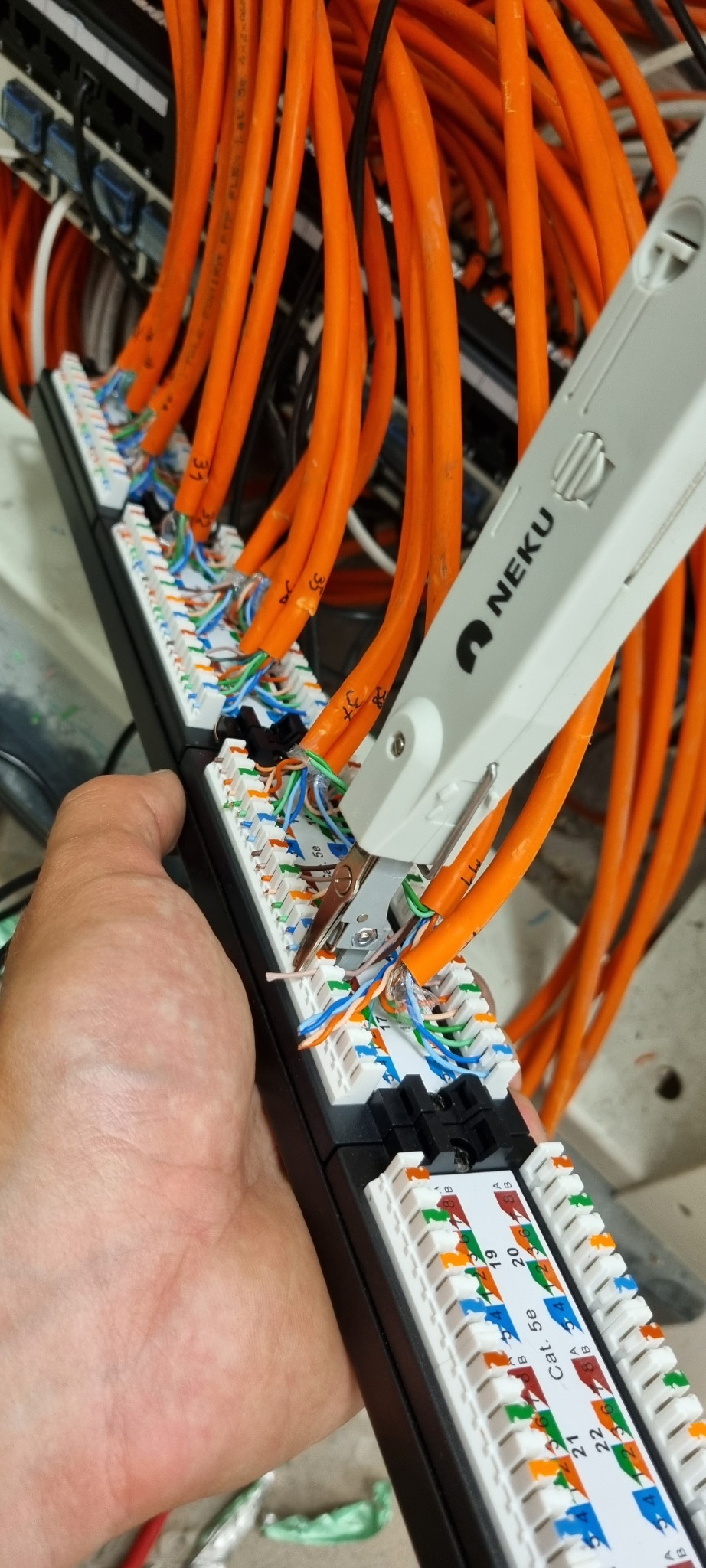 Ręczne zarabianie pomarańczowych kabli sieciowych w panelu krosowniczym Cat 5e przy użyciu narzędzia NEKU, widoczne oznaczenia numerów portów i żyły kabli w kolorach.