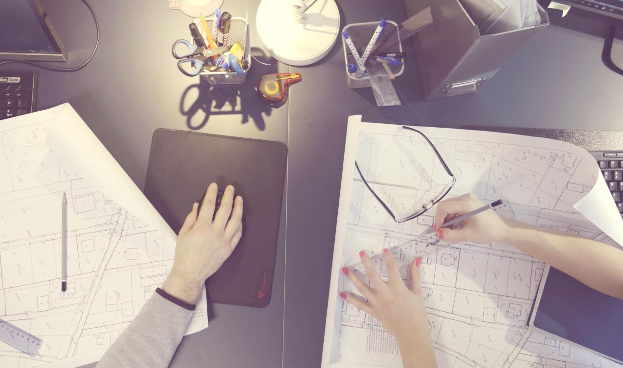 Biuro projektowe: Dwie osoby pracują nad planami na stole, jedna ręka na myszce, druga precyzyjnie rysuje ołówkiem po planie, używając linijki i okularów.