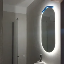 Nowoczesna łazienka z owalnym lustrem podświetlanym LED, na którym wciąż znajduje się niebieska folia ochronna, minimalistyczna umywalka z baterią, szklana kabina prysznicowa w tle, jasne płytki...