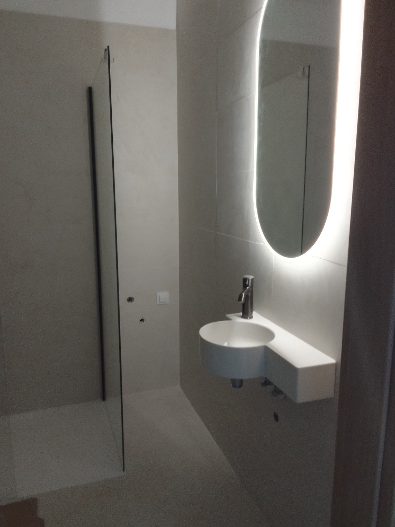 Minimalistyczna łazienka w odcieniach bieli z owalnym lustrem podświetlonym LED, wiszącą umywalką i kabiną prysznicową typu walk-in.