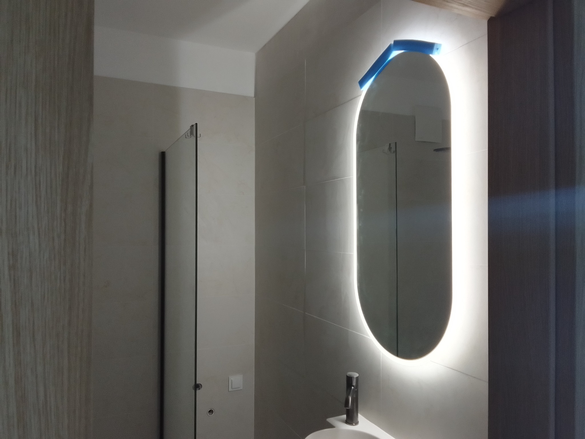 Nowoczesna łazienka z owalnym lustrem podświetlanym LED, na którym wciąż znajduje się niebieska folia ochronna, minimalistyczna umywalka z baterią, szklana kabina prysznicowa w tle, jasne płytki...