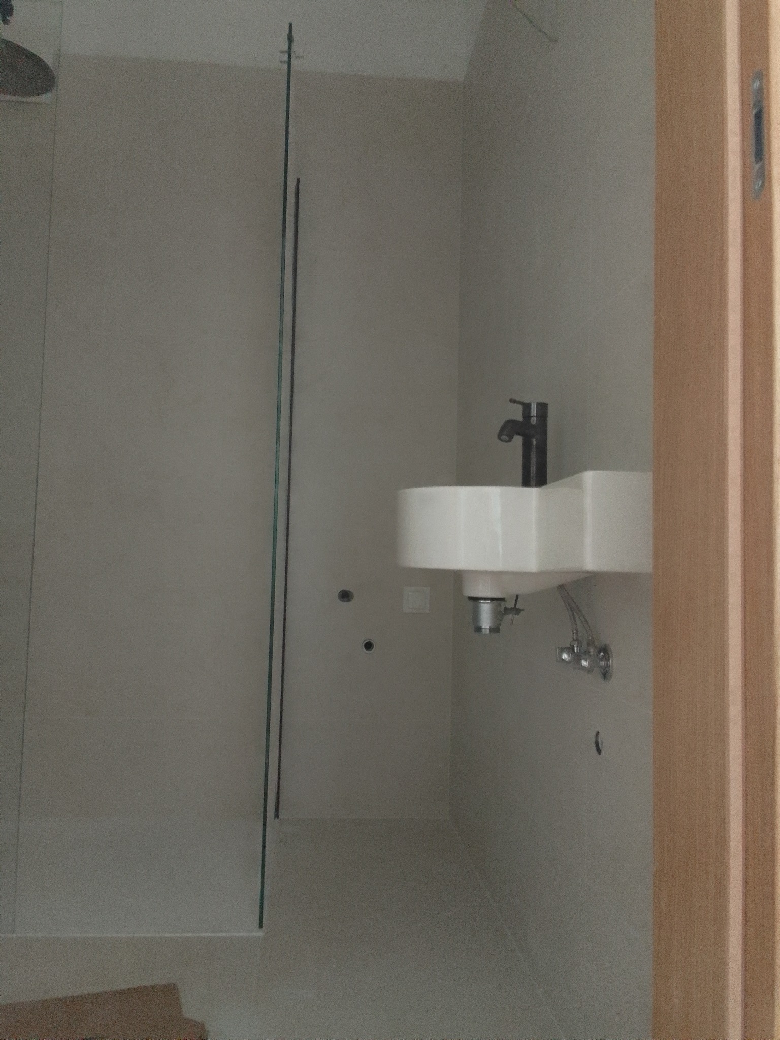 Nowoczesna łazienka z kabiną prysznicową oddzieloną szklaną ścianką, białą umywalką na ścianie i czarną baterią, widok zza drewnianej futryny drzwi.