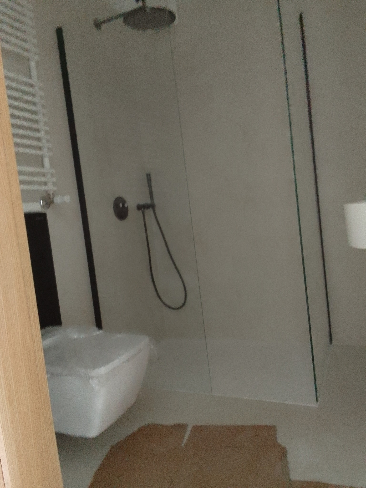 Nowoczesna, jasna łazienka w trakcie wykańczania, z kabiną prysznicową typu walk-in z czarnymi ramami, podwieszaną toaletą owiniętą folią ochronną i grzejnikiem drabinkowym.