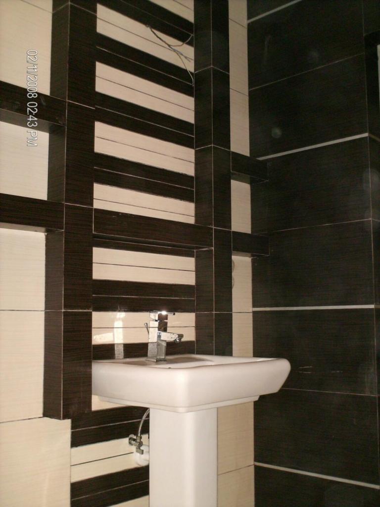 Umywalka na postumencie z chromowaną baterią, otoczona ścianami wyłożonymi płytkami w odcieniach brązu i beżu, z widocznymi przewodami na górze.