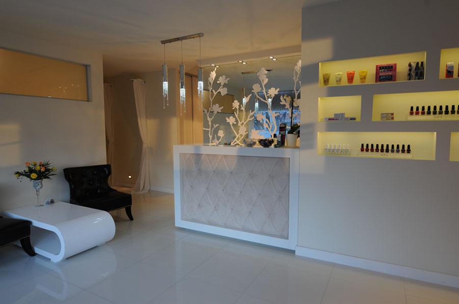 Eleganckie wnętrze salonu kosmetycznego z recepcją z pikowanym frontem, szklaną ścianką z motywem kwiatowym, wiszącymi lampami kryształowymi i półkami z lakierami do paznokci w podświetlanych niszach.
