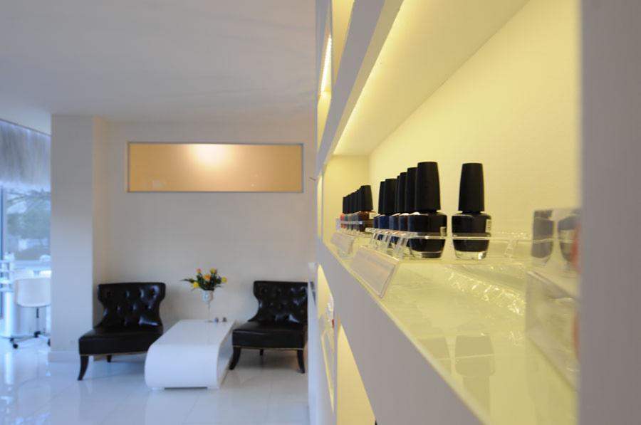 Wnętrze salonu kosmetycznego: rzędy lakierów do paznokci na podświetlonej półce, w tle stolik z kwiatami i czarne fotele.