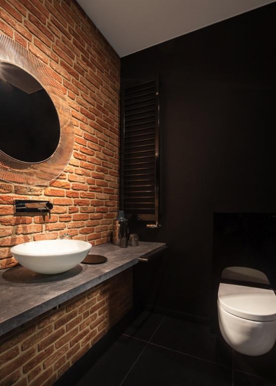 Nowoczesna łazienka z czerwoną ścianą z cegły, okrągłym lustrem z podświetleniem, białą umywalką nablatową i czarną ścianą oraz grzejnikiem drabinkowym.